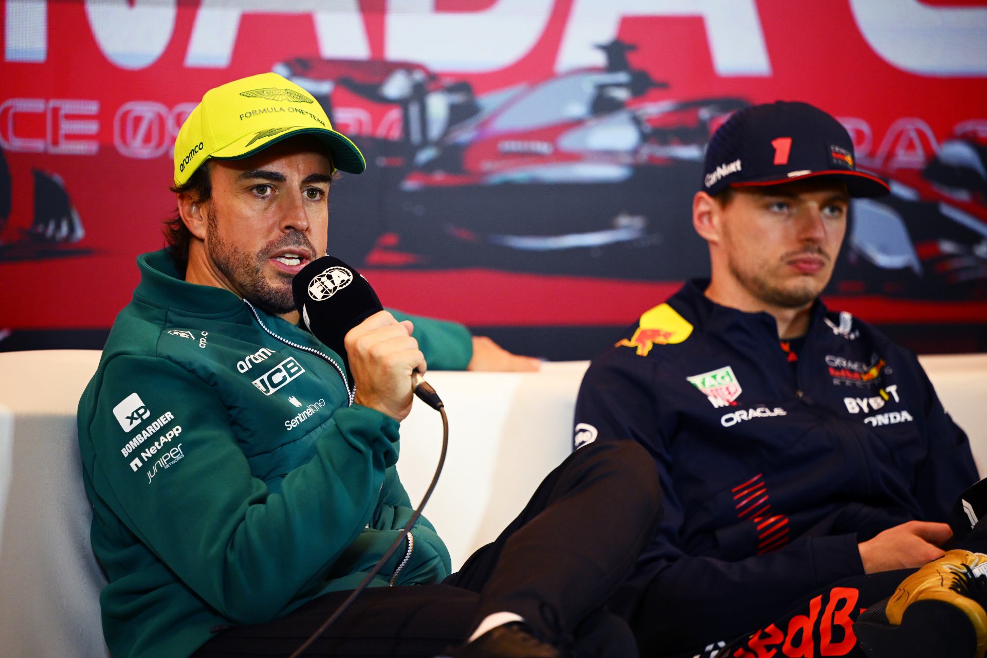 Fernando Alonso talking at a press conference with Max Verstappen in the background